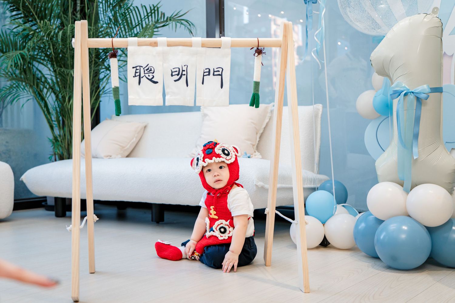 A nursery decorated with blue balloons features a wooden clothing rack and white sofa against a cloudy wallpaper background.