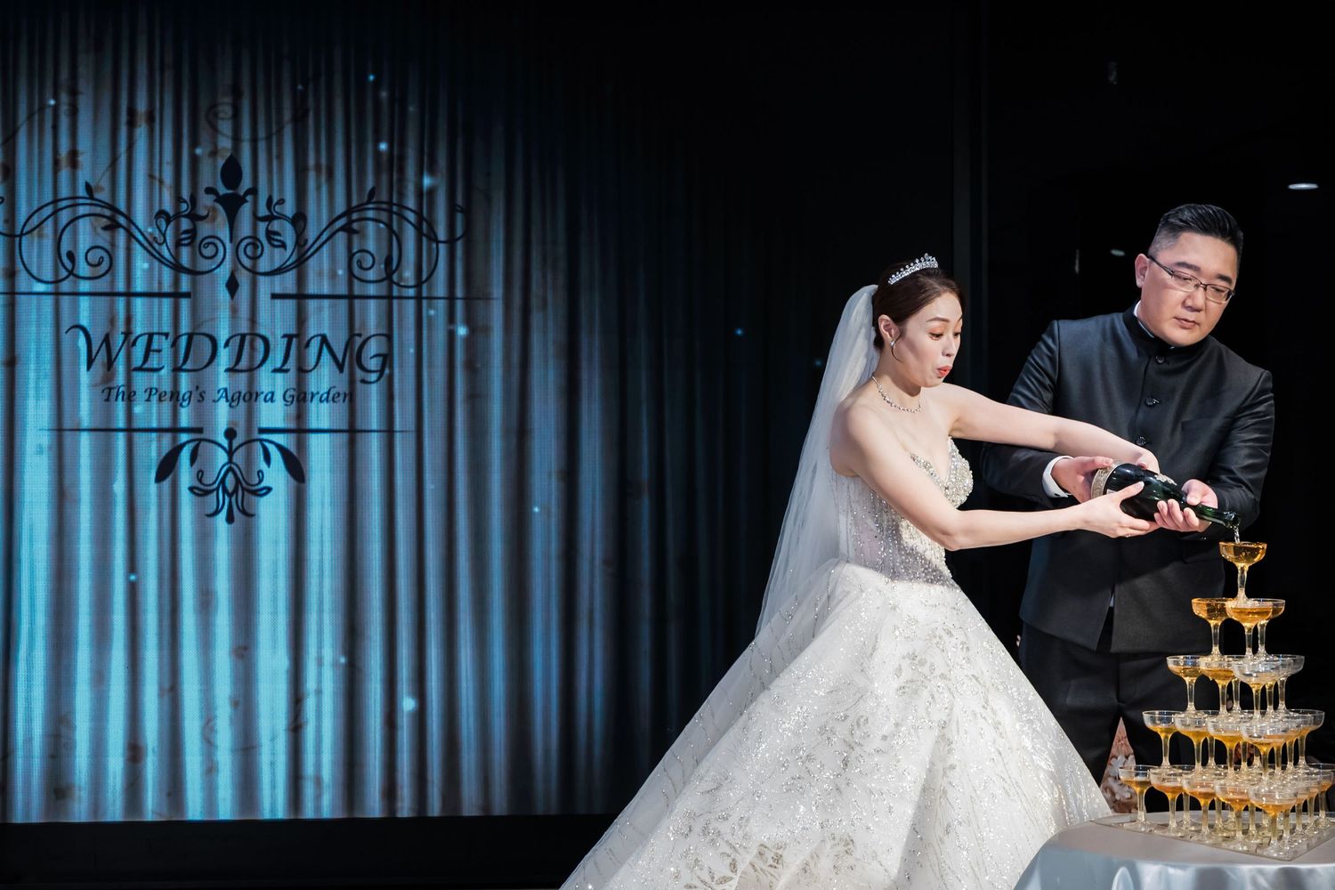 Bride and groom pour champagne into a tower of glasses at their wedding reception with blue lighting.