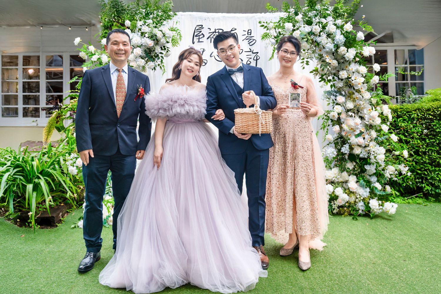 Four people pose together under a floral arch at a formal celebration event on green grass.