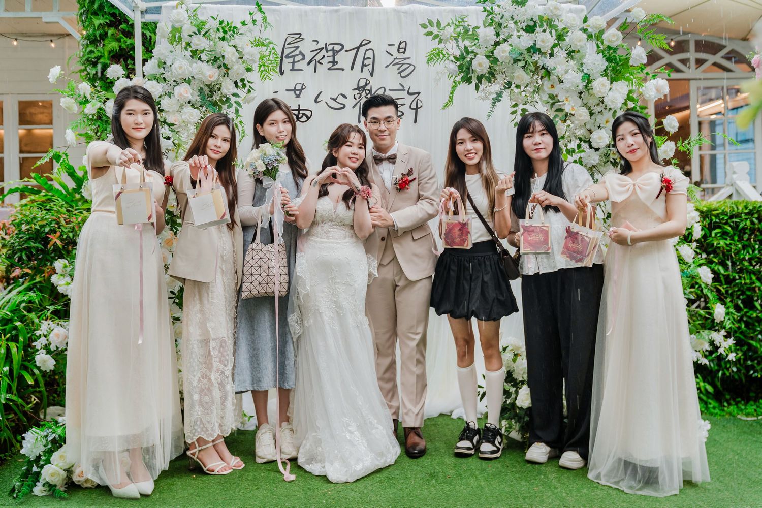 A group in pastel floral dresses and casual attire poses together at a garden-themed event with white floral decorations.