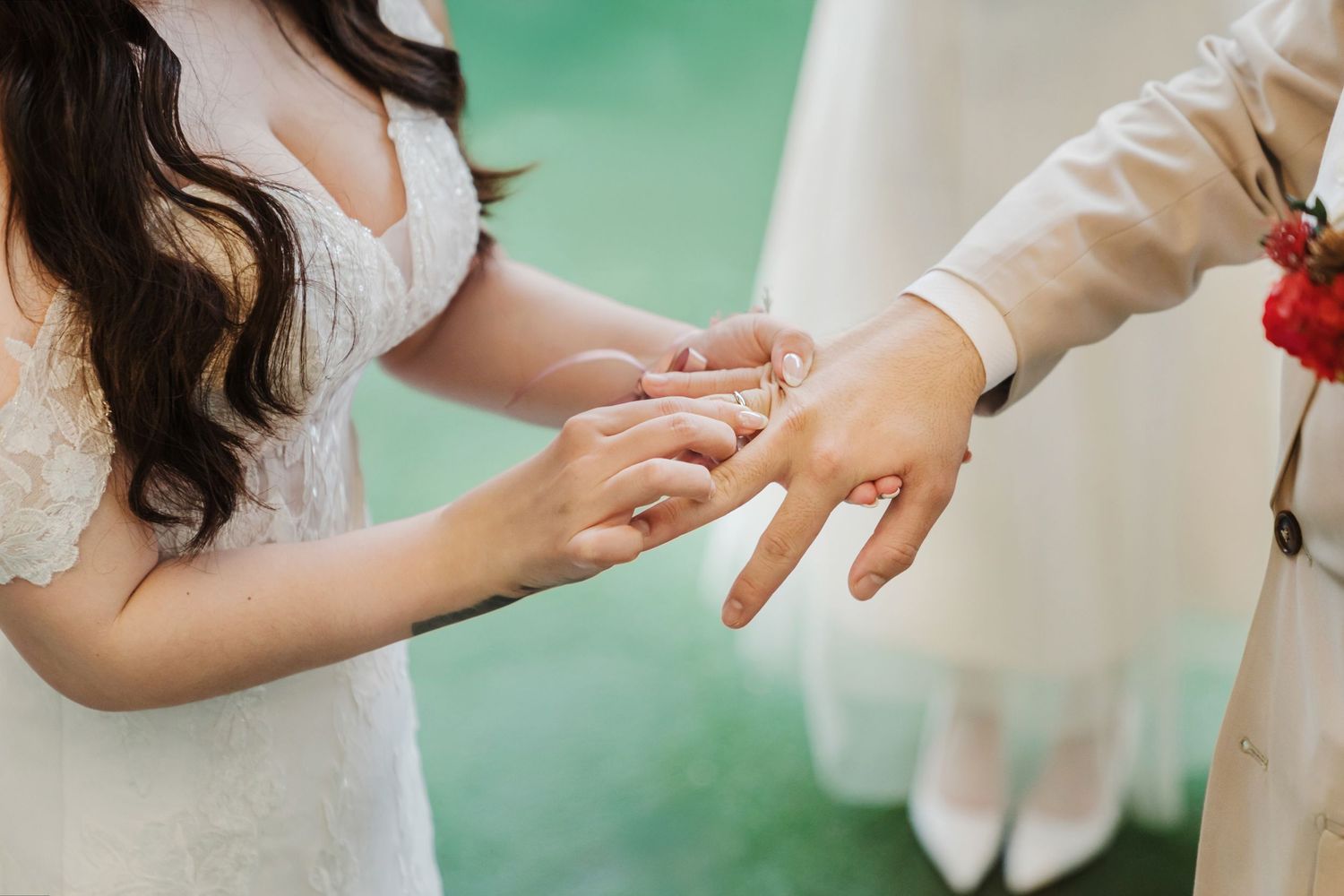 Close up of hands as someone places wedding ring on finger during marriage ceremony against blurred green background.
