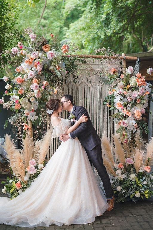 A romantic wedding scene with a floral macrame arch decorated with pink and peach roses, pampas grass and greenery outdoors.