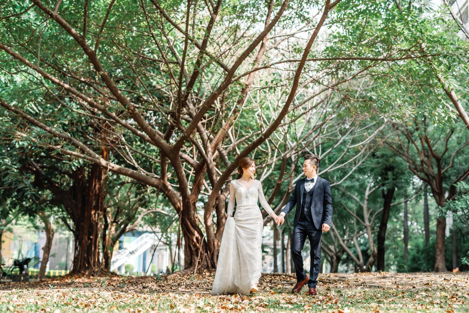 A couple walks hand in hand through a lush garden path beneath sprawling trees on their wedding day.