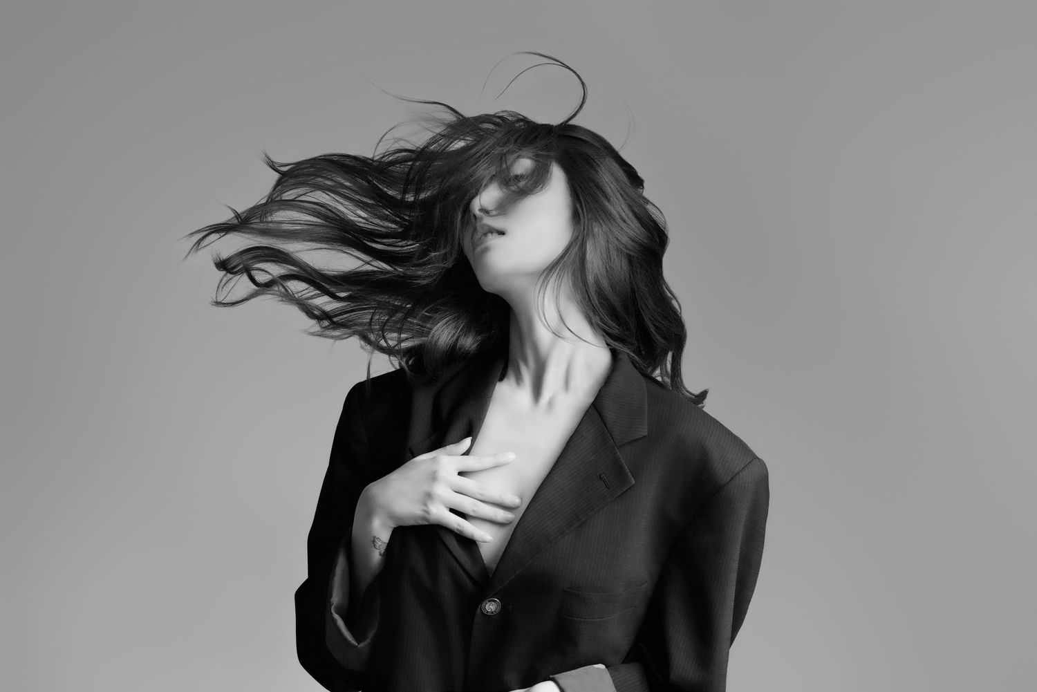 A figure in black jacket flips their dark hair in dramatic motion against a gray studio backdrop.