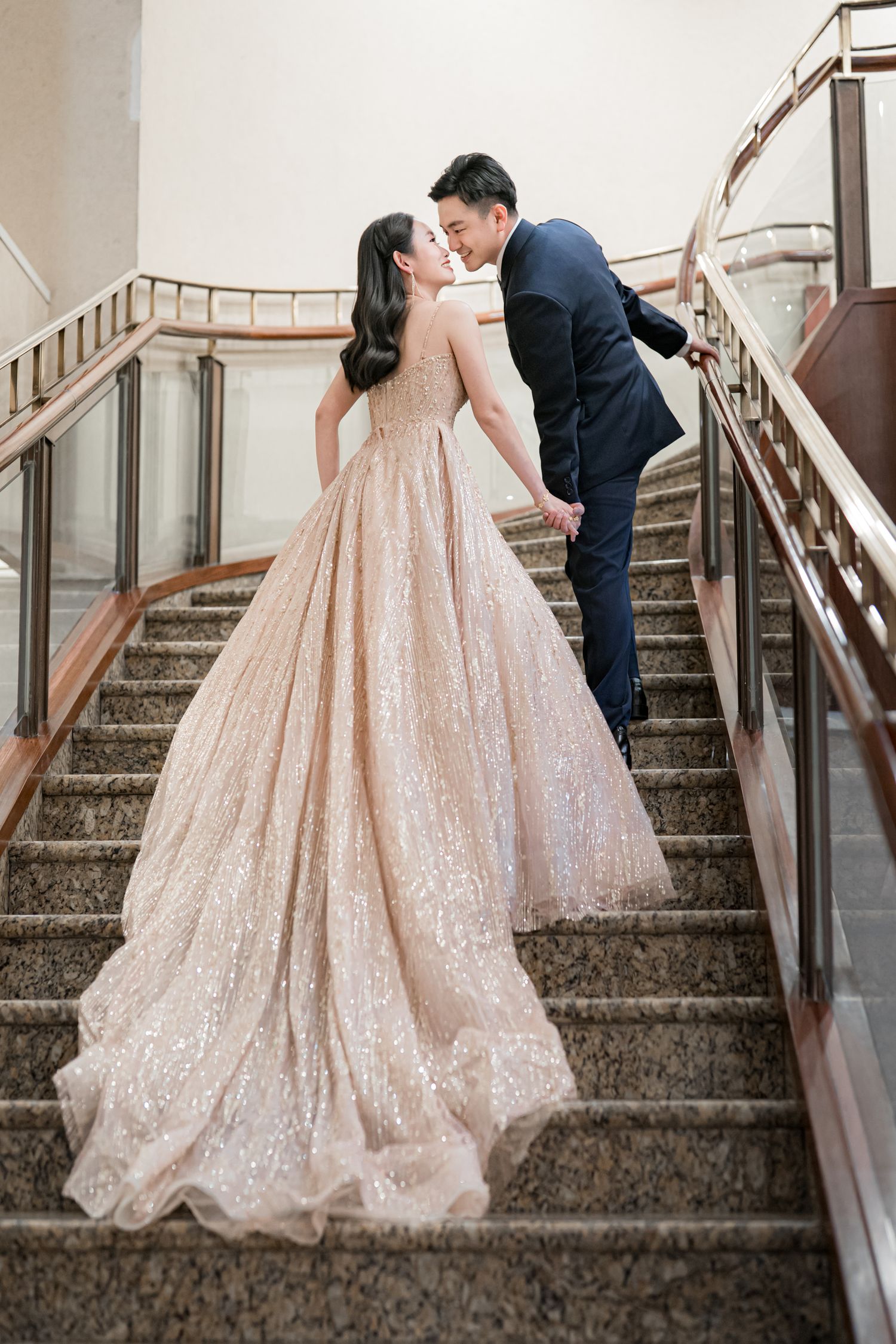 A couple shares a romantic kiss on a grand staircase, with the bride wearing a sparkling blush ballgown.