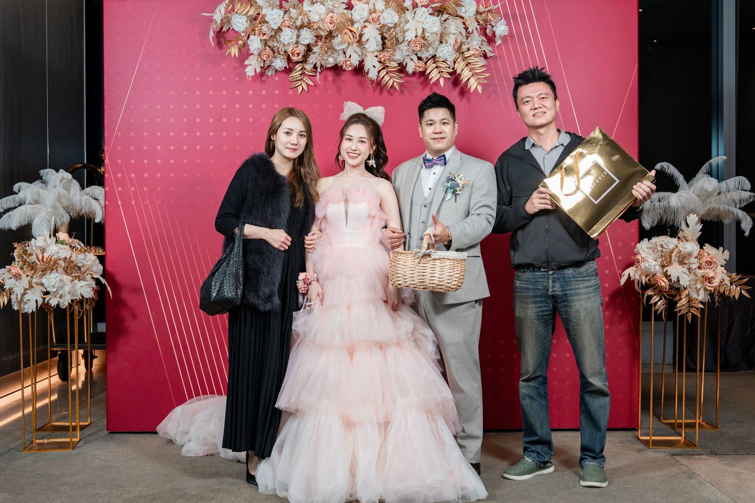 Group gathers for celebratory photo against pink wall with elegant white floral arrangement overhead.