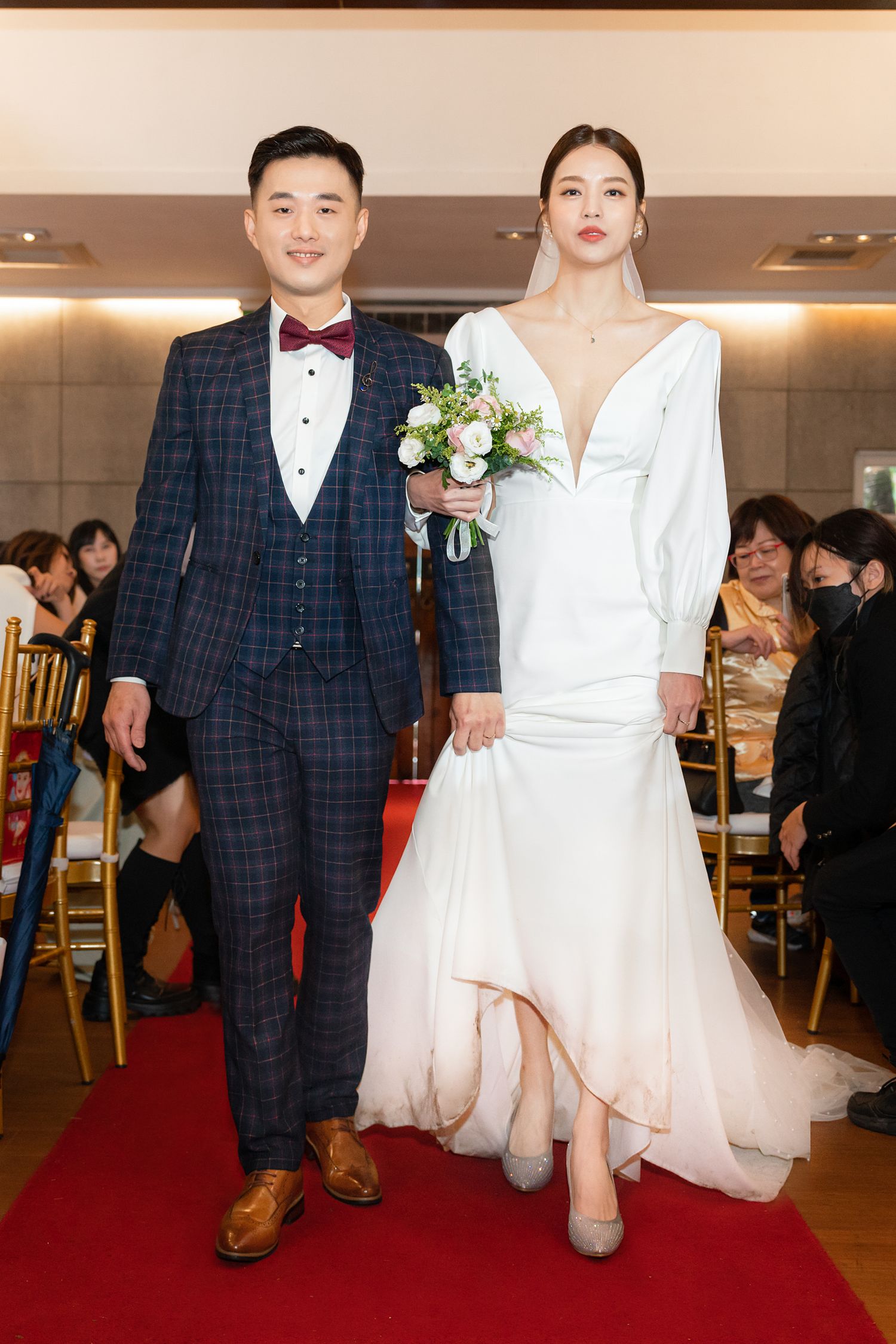 A couple walks down a red carpet at a wedding ceremony, with the groom in plaid suit and bride in elegant white gown.