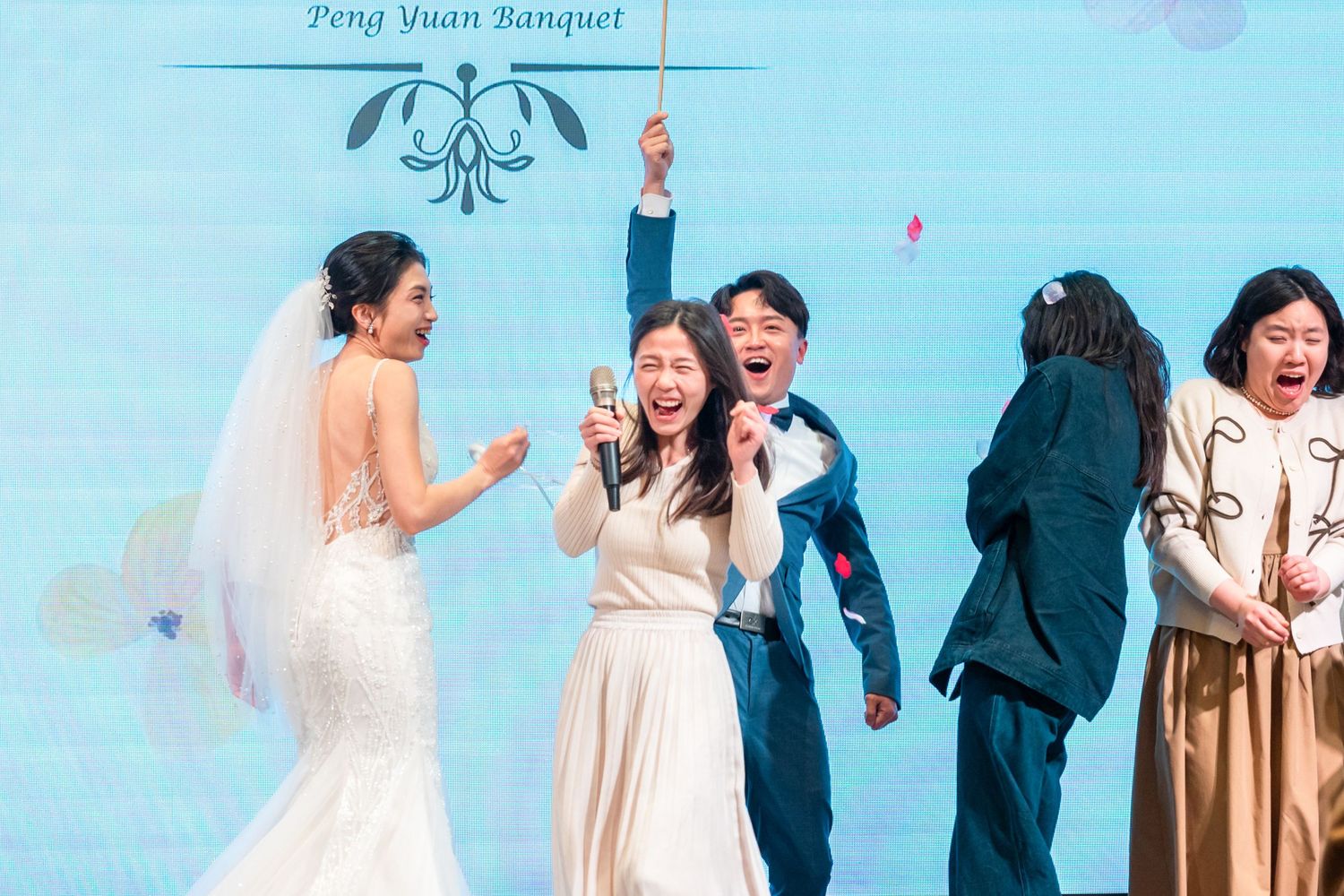 Wedding celebration scene with joyful dancing and reactions during reception under blue lighting and floral decor.