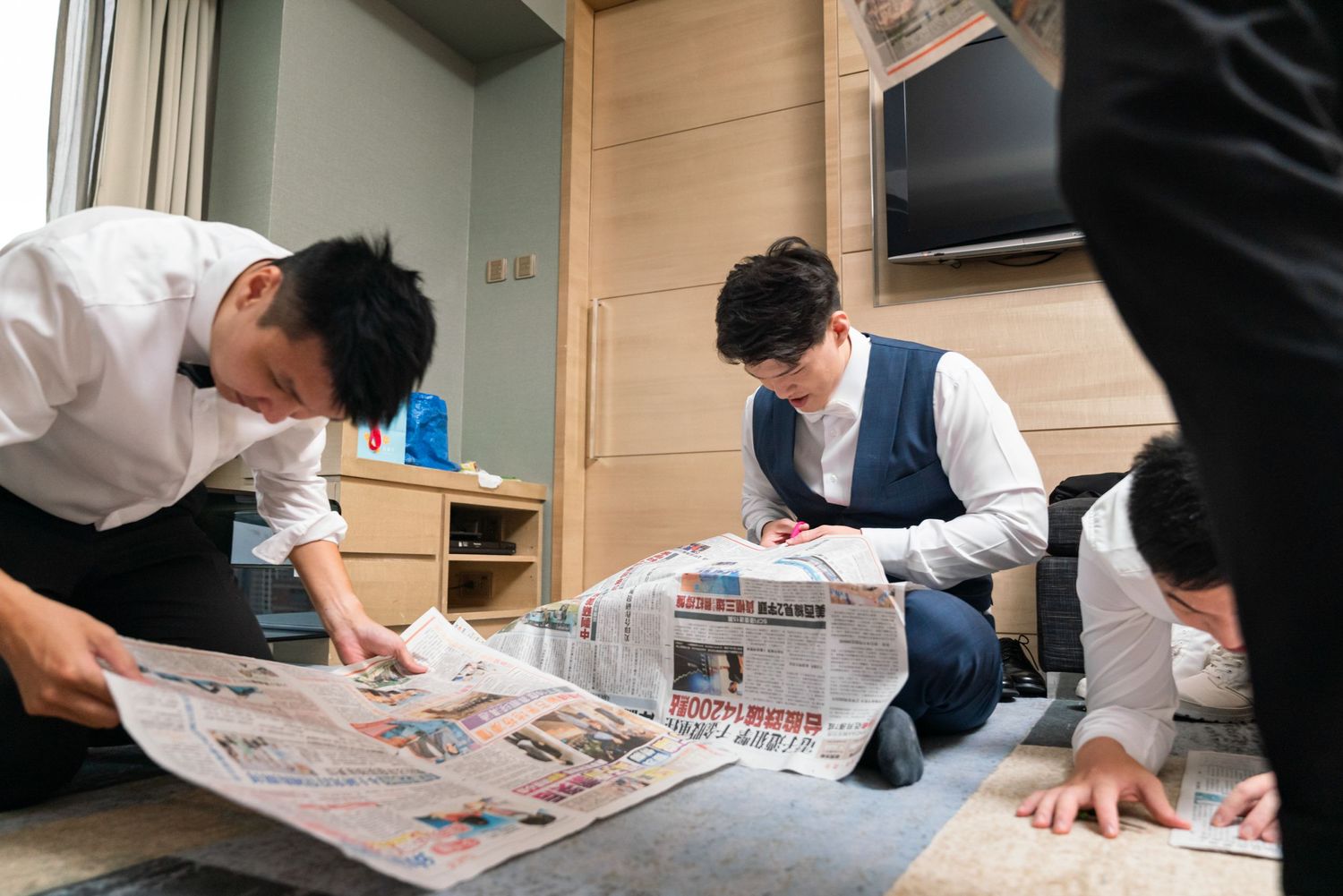 Three people wearing face masks sit in a room reading through newspapers spread out on the floor.