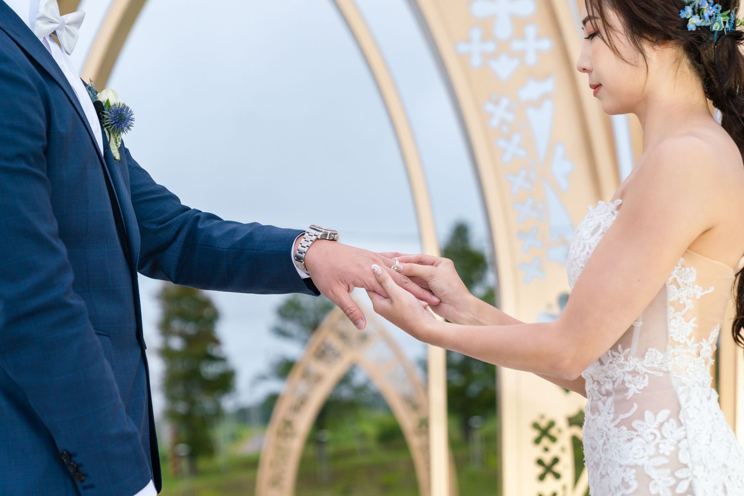 Closeup of couple holding hands during outdoor wedding ceremony under decorative arch with floral details.