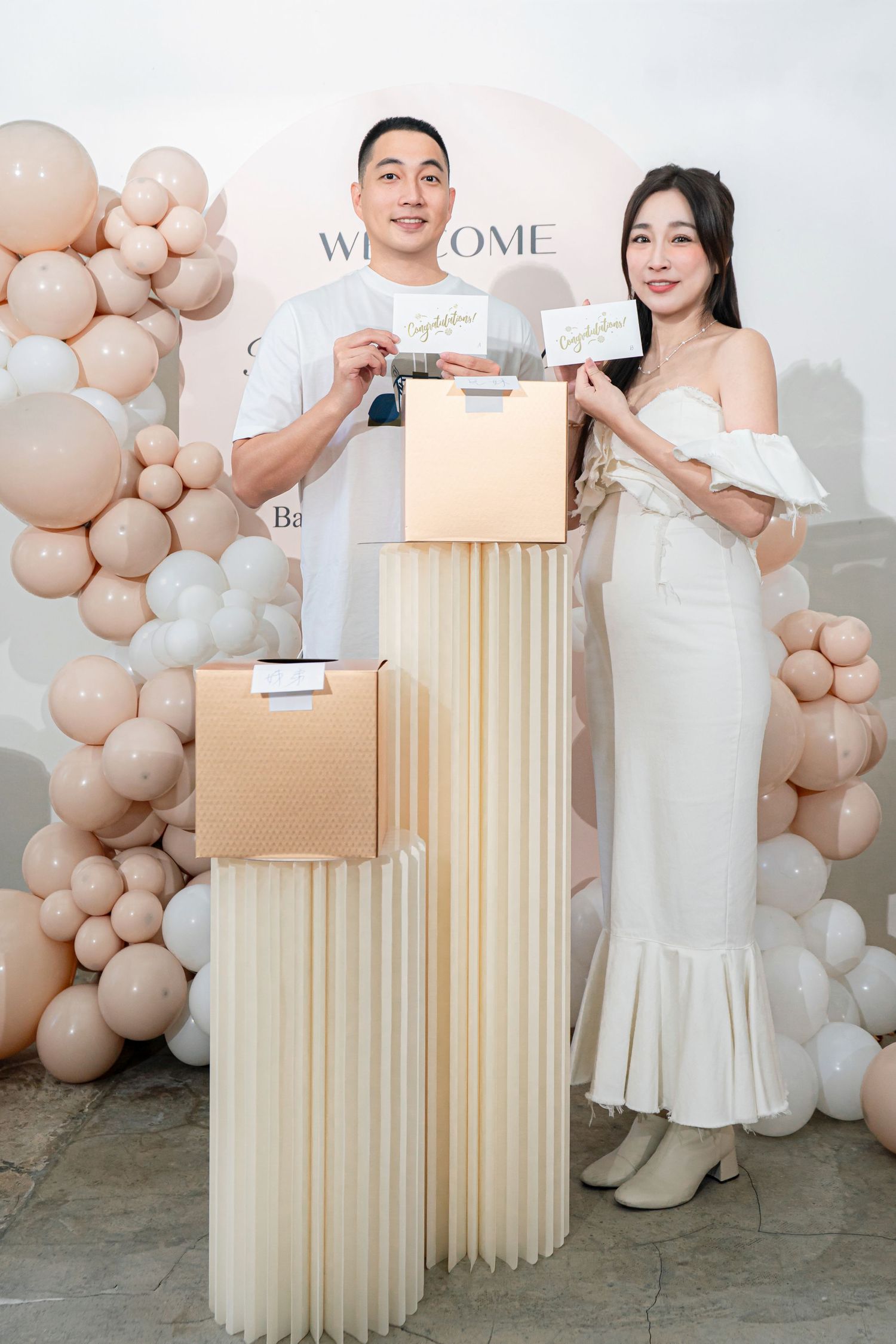 A couple poses with gift boxes at an elegant event setup with beige balloon arch and decorative columns.