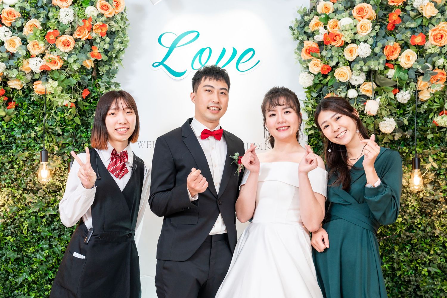 Wedding photo with couple and guests in front of floral wall display with blue 'Love' text.