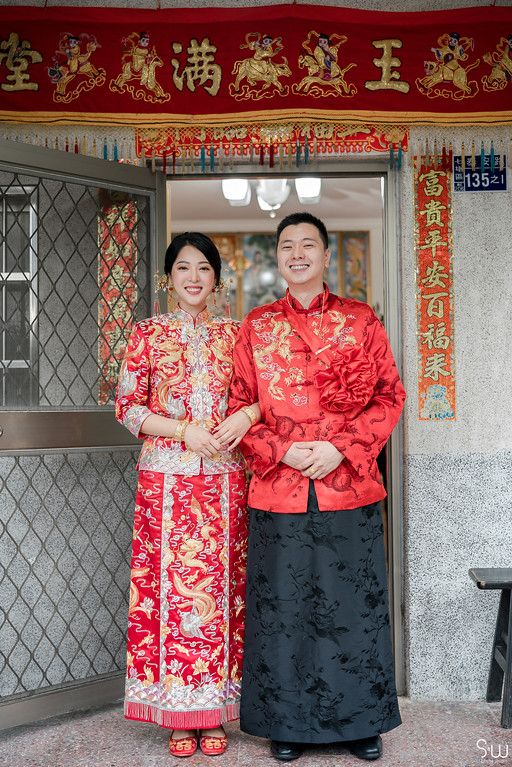 Couple poses in traditional Chinese wedding attire under red decorative banners.