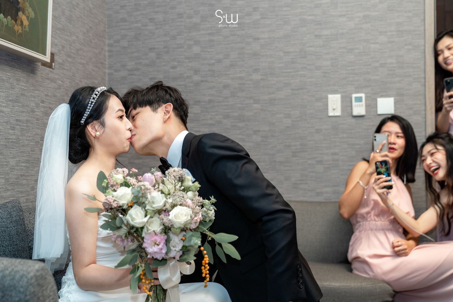 A wedding couple shares a kiss while guests capture the moment on their phones in the background.