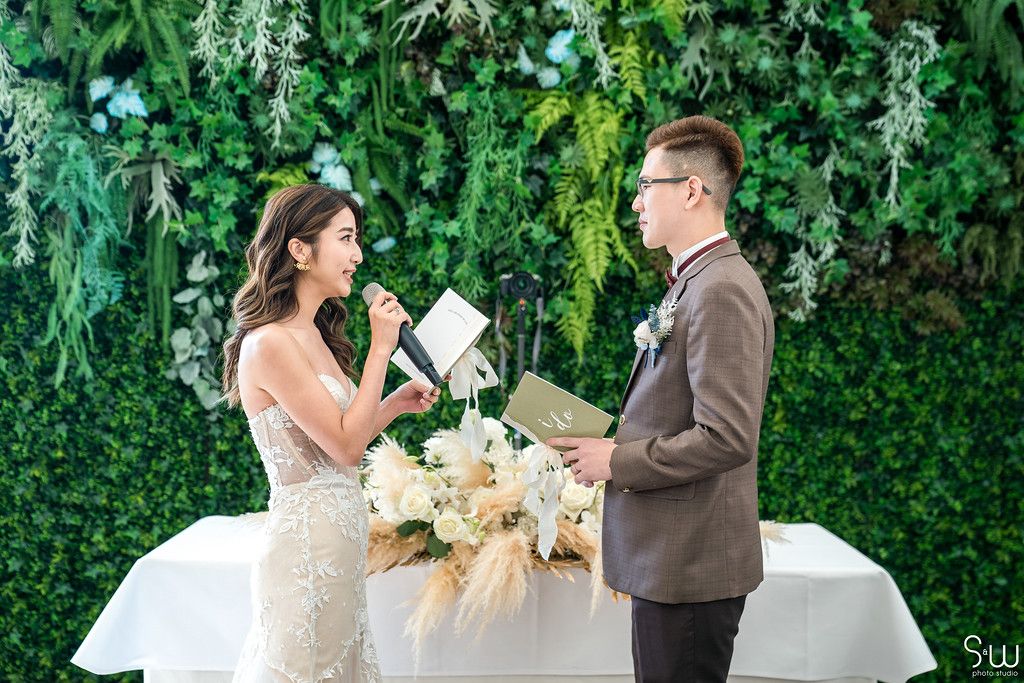 Couple shares heartfelt vow exchange during outdoor ceremony with floral wall backdrop.