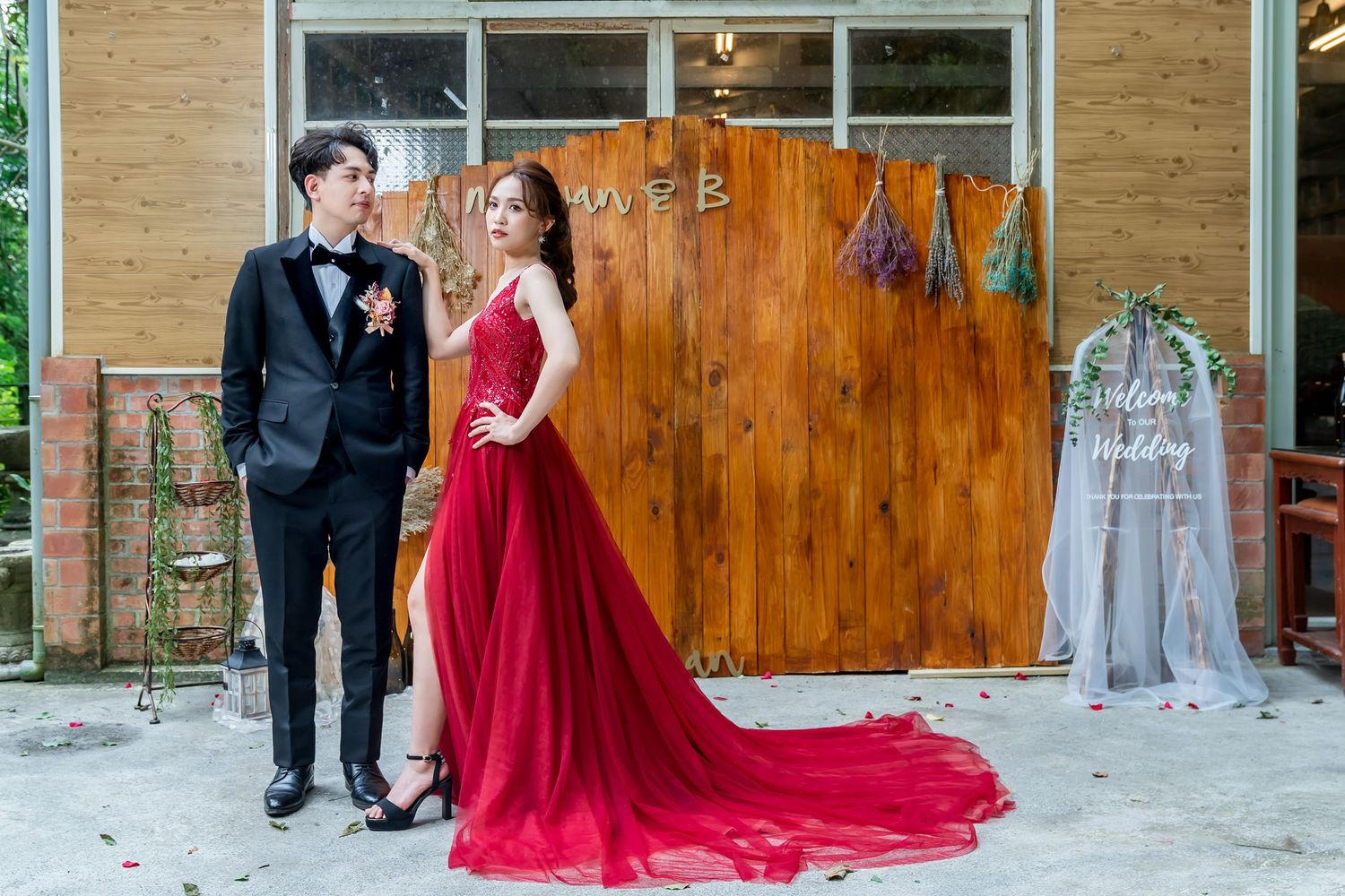 Formal couple pose in front of rustic wooden wall with red flowing gown and classic tuxedo.