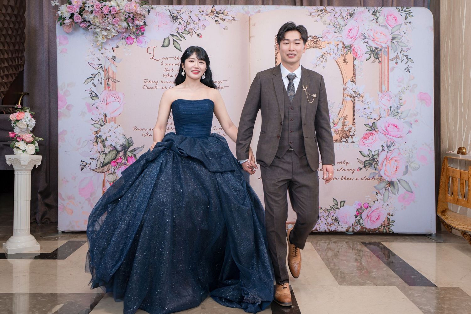 Couple posing at wedding reception, woman in navy blue ballgown and man in gray suit, floral backdrop.