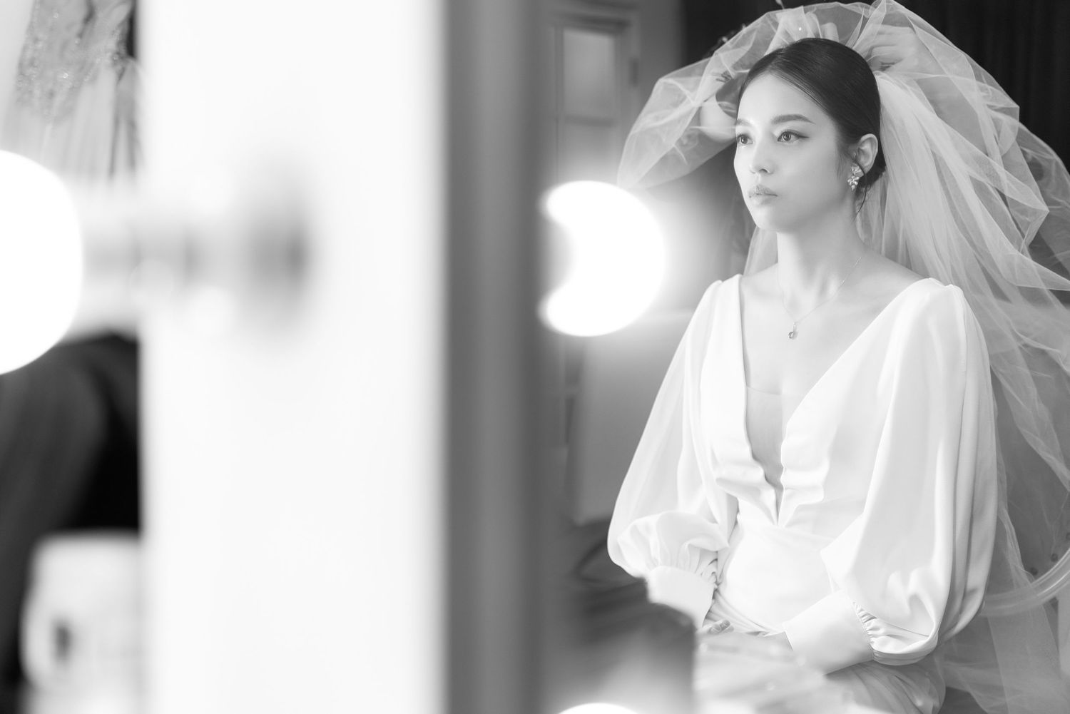 A bride in a white wedding dress with billowing sleeves and veil stands in soft black and white lighting.