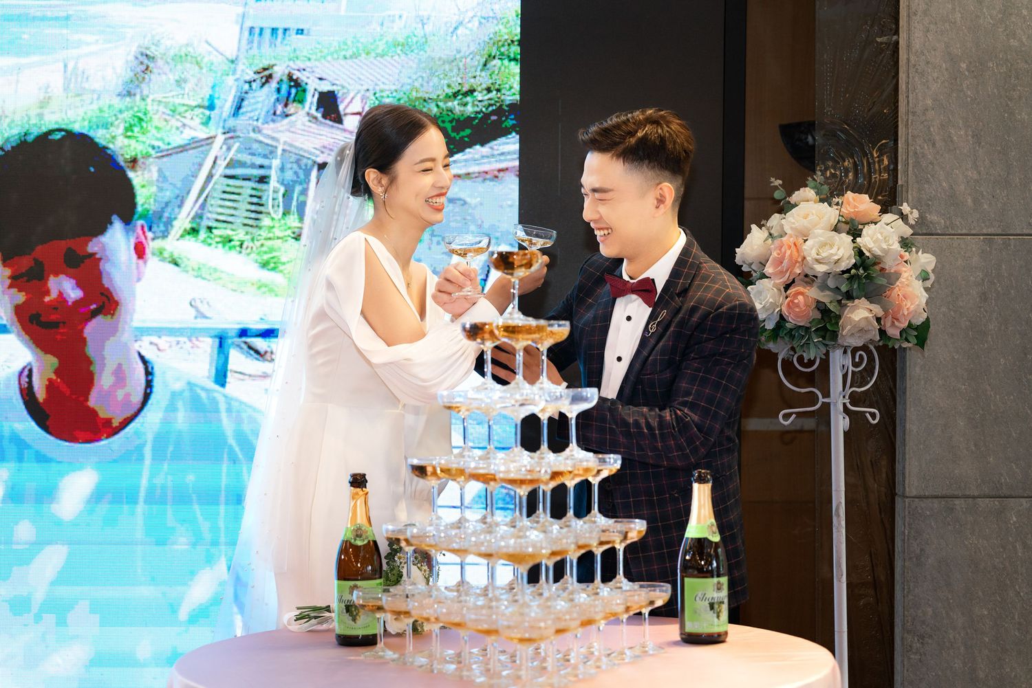 A couple pours champagne into a tower of glasses at an elegant wedding celebration with floral decorations.
