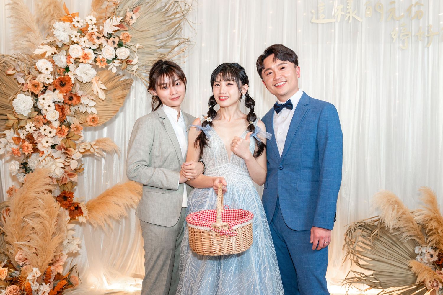 Three people pose together at a wedding celebration with floral decorations and a pink-filled basket.