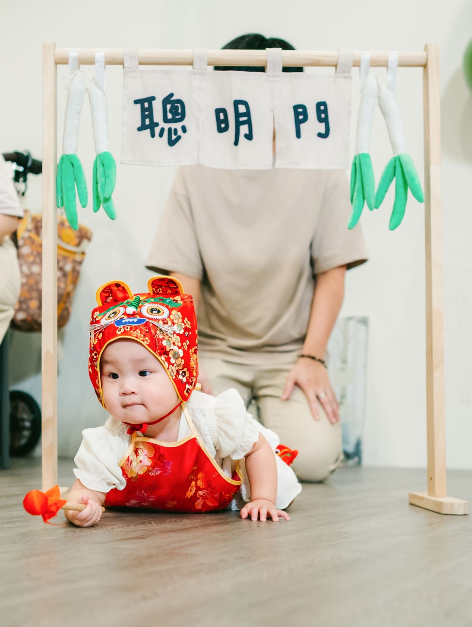 Baby wearing traditional red Chinese New Year outfit with tiger hat crawls on wooden floor while adult stands nearby.