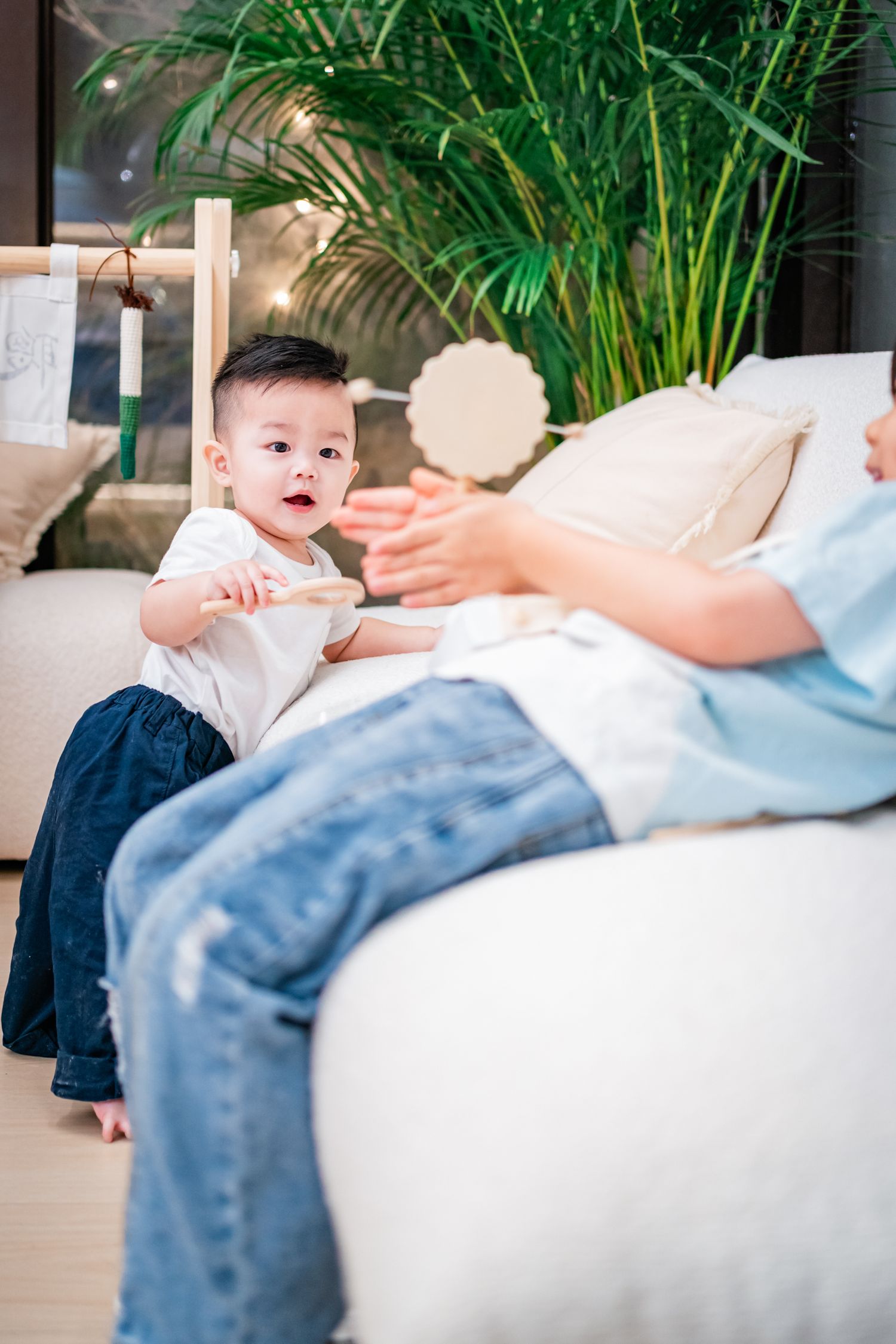 A toddler in blue shorts sits on a white couch while an adult hand reaches toward them in a bright indoor setting.