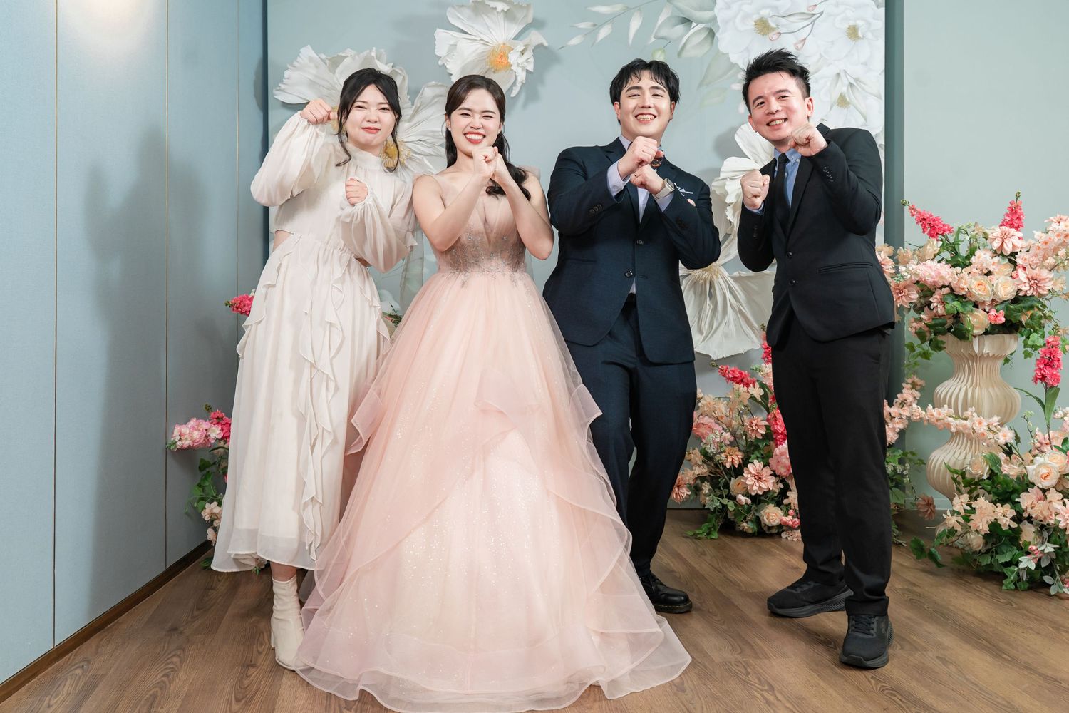 Wedding party poses together in formal attire with pink floral decorations against blue wall backdrop.