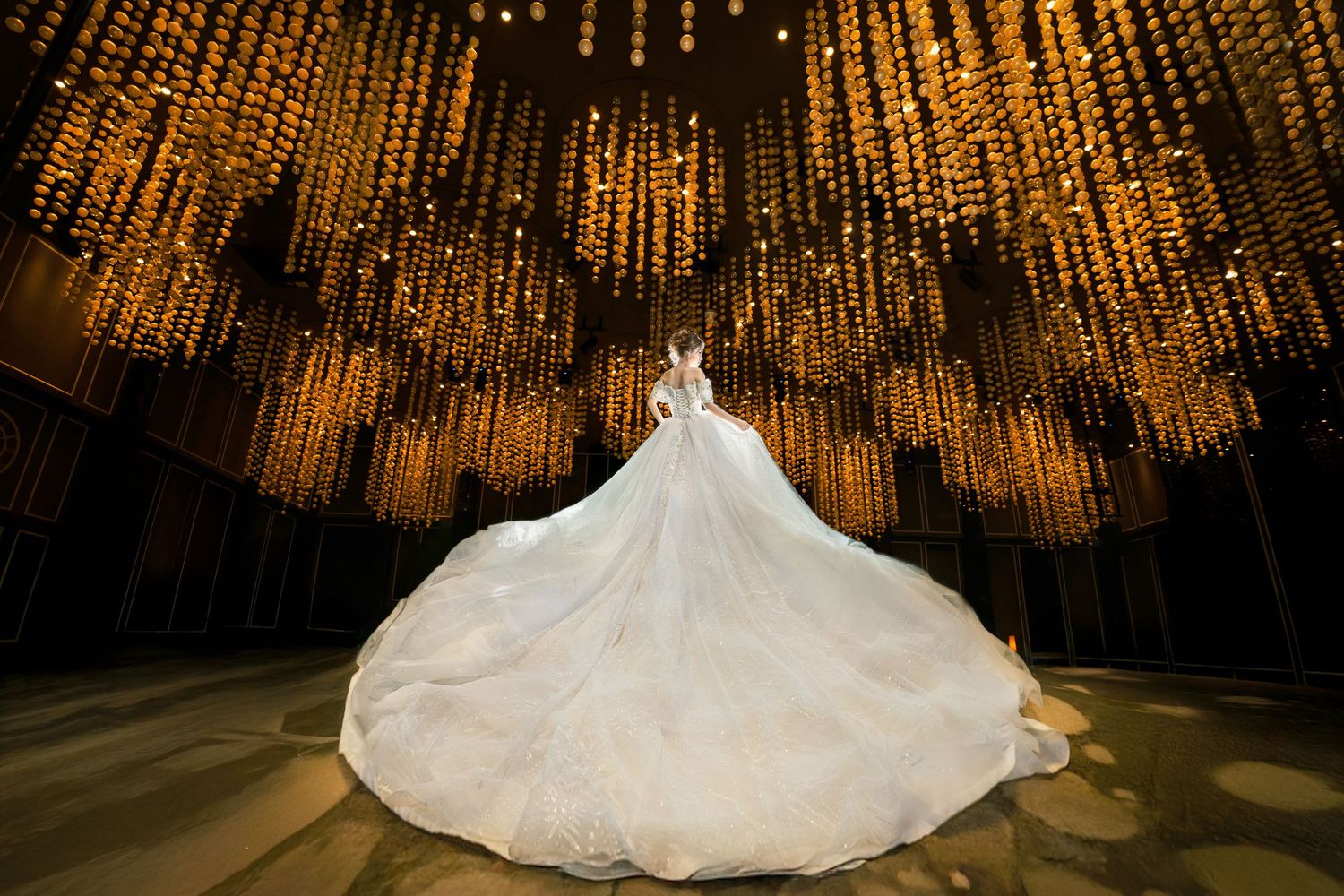 A bride in a flowing white ballgown stands beneath a stunning ceiling of glowing golden string lights.