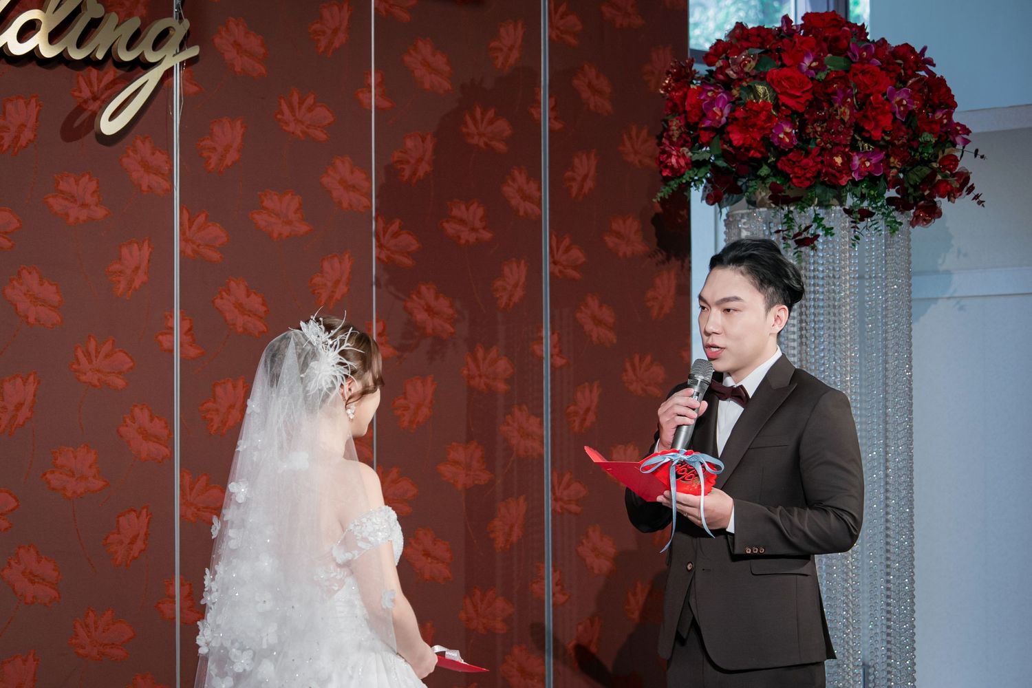 Wedding couple poses against red floral wallpaper backdrop in elegant formal attire.