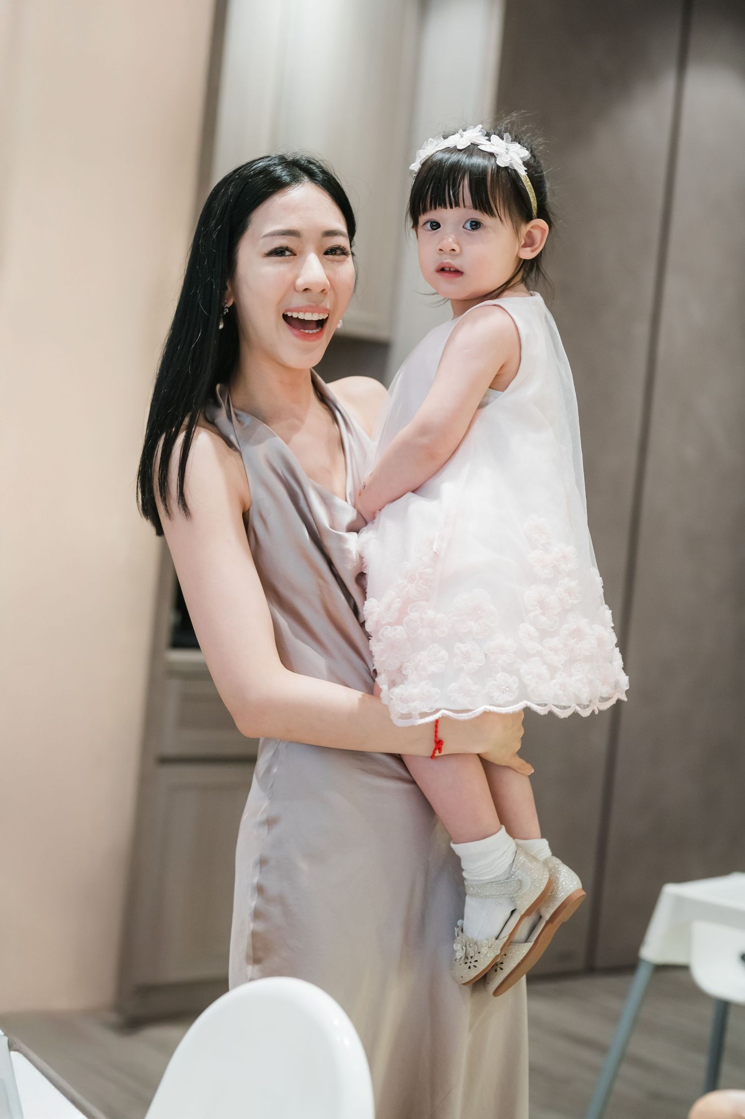 A joyful mother in a gray dress lifts and plays with her baby wearing a white dress against a neutral background.