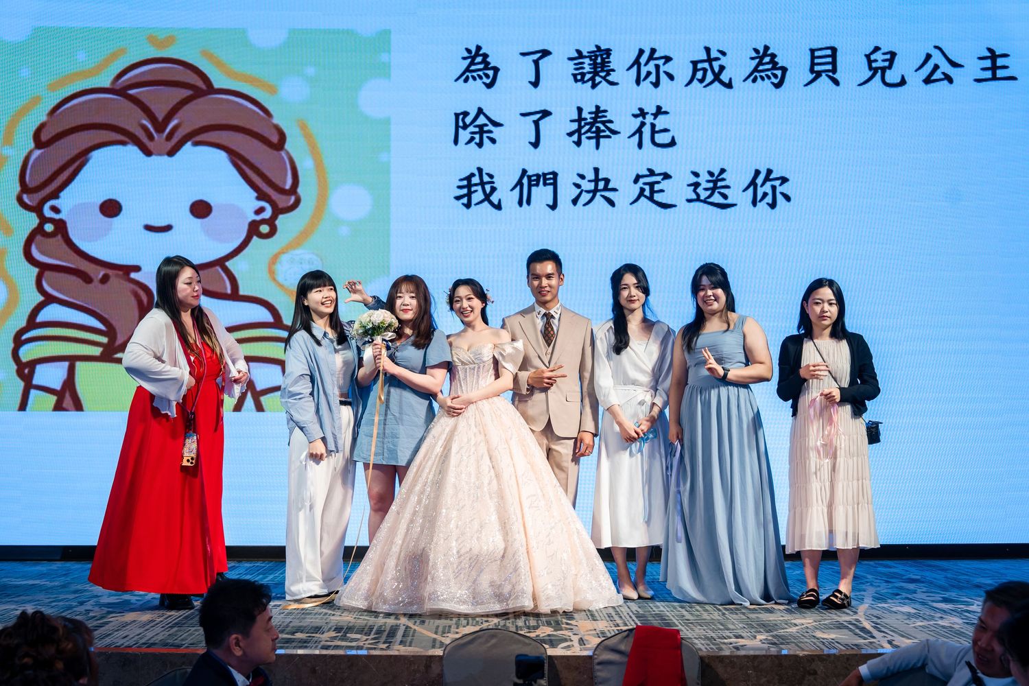 Wedding group photo on stage with bride in white gown and guests holding bouquet at event.
