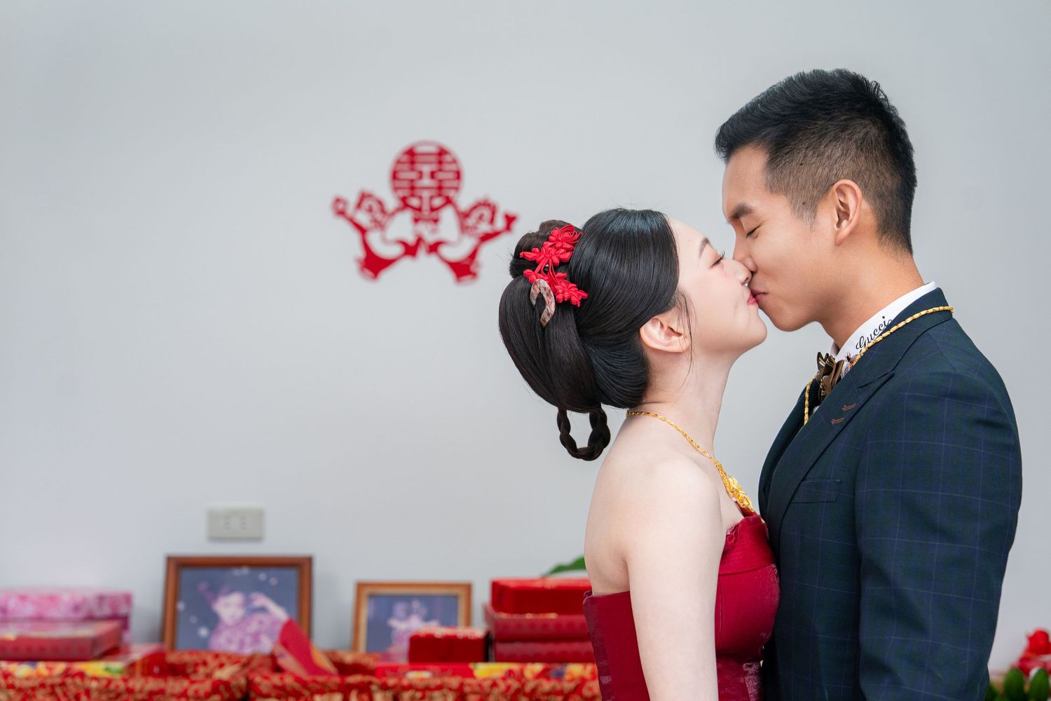 Chinese wedding couple sharing a kiss with traditional red double happiness symbol on wall behind them.