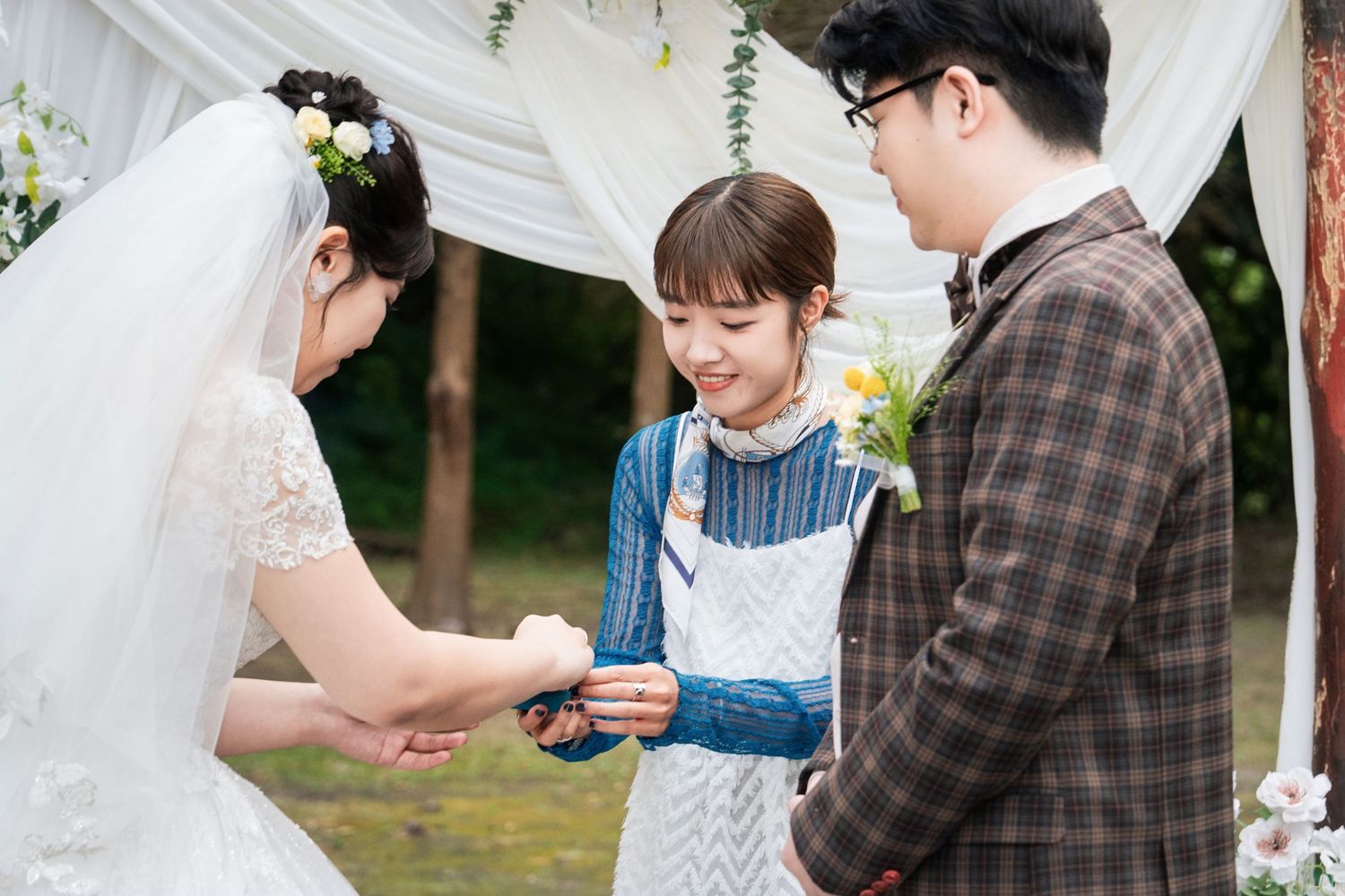 A bride and groom exchange rings during an outdoor garden wedding ceremony beneath a white arch.