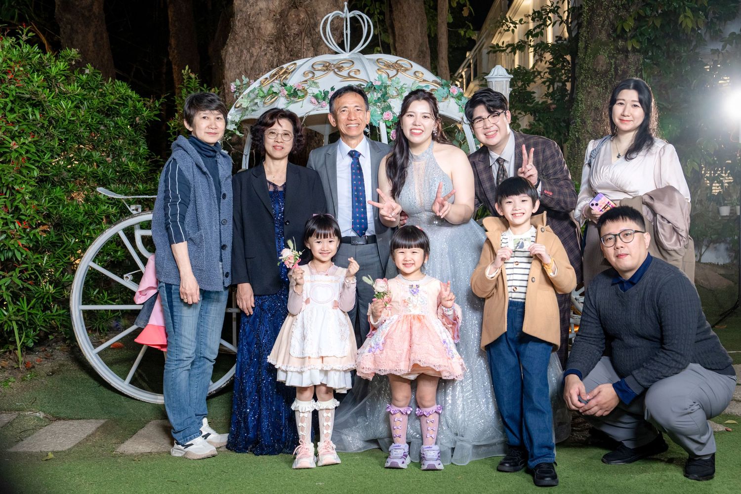 A group photo of people standing together at an outdoor garden venue with decorative lighting and a vintage wheel prop.