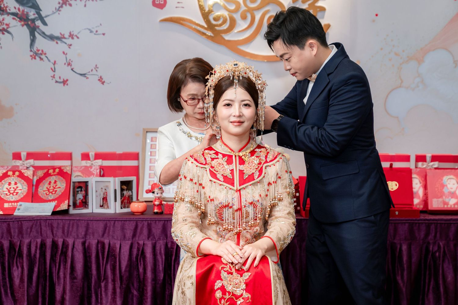 A bride in a red and gold Chinese wedding gown and phoenix headpiece is helped by a groom and a woman during a traditional tea ceremony.