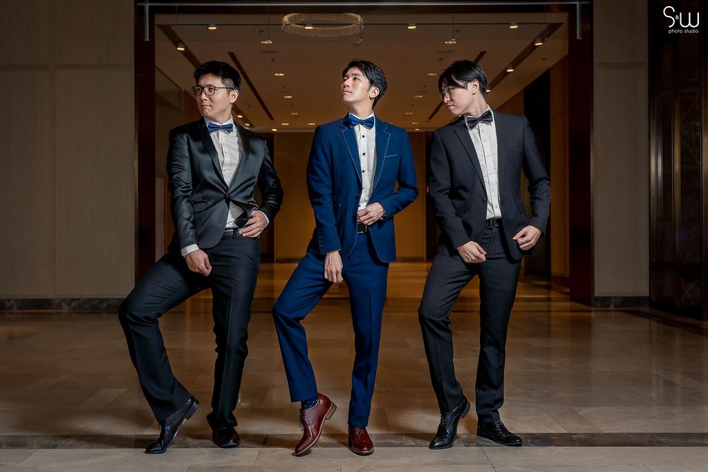 Three groomsmen in formal suits pose together against a wall in coordinated stances.