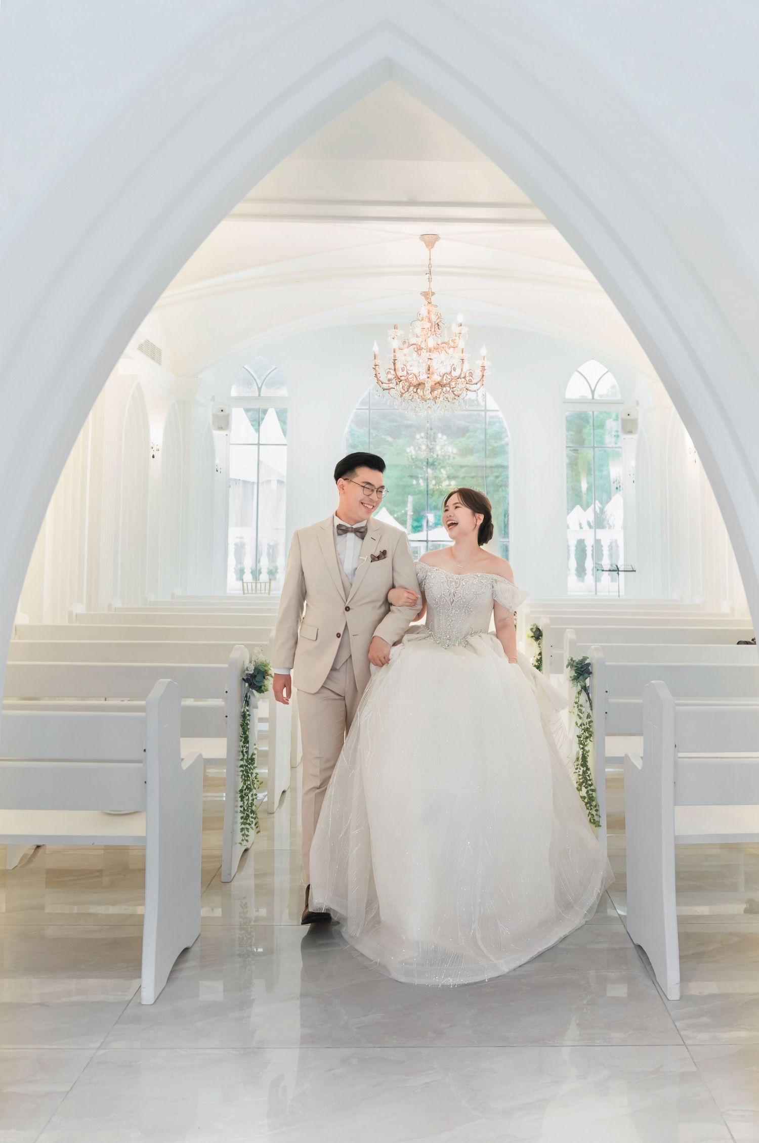 Elegant wedding couple standing under an archway in a bright white chapel with a crystal chandelier.