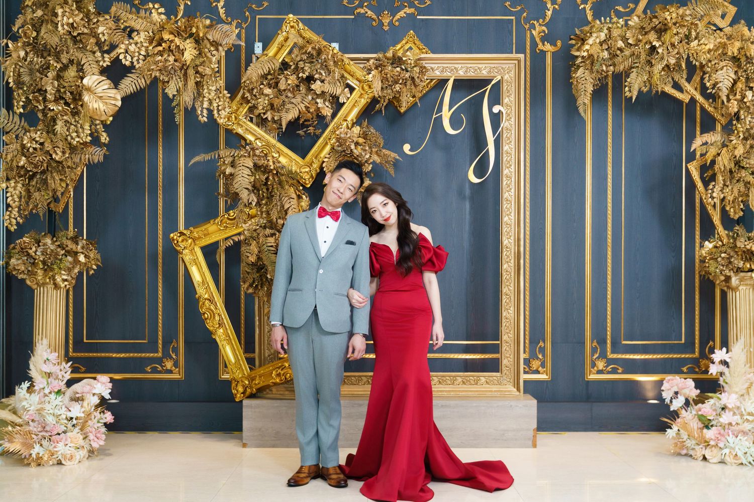 A couple poses against an ornate navy blue wall with golden frames and floral decorations at a formal event.