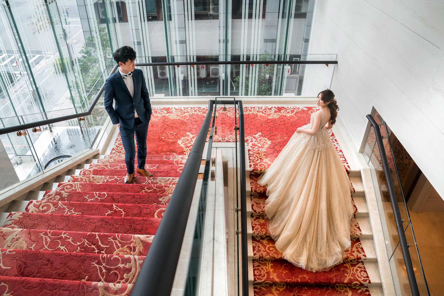 Two people stand on opposite sides of an elegant red-carpeted staircase with a glass railing in a modern interior space.