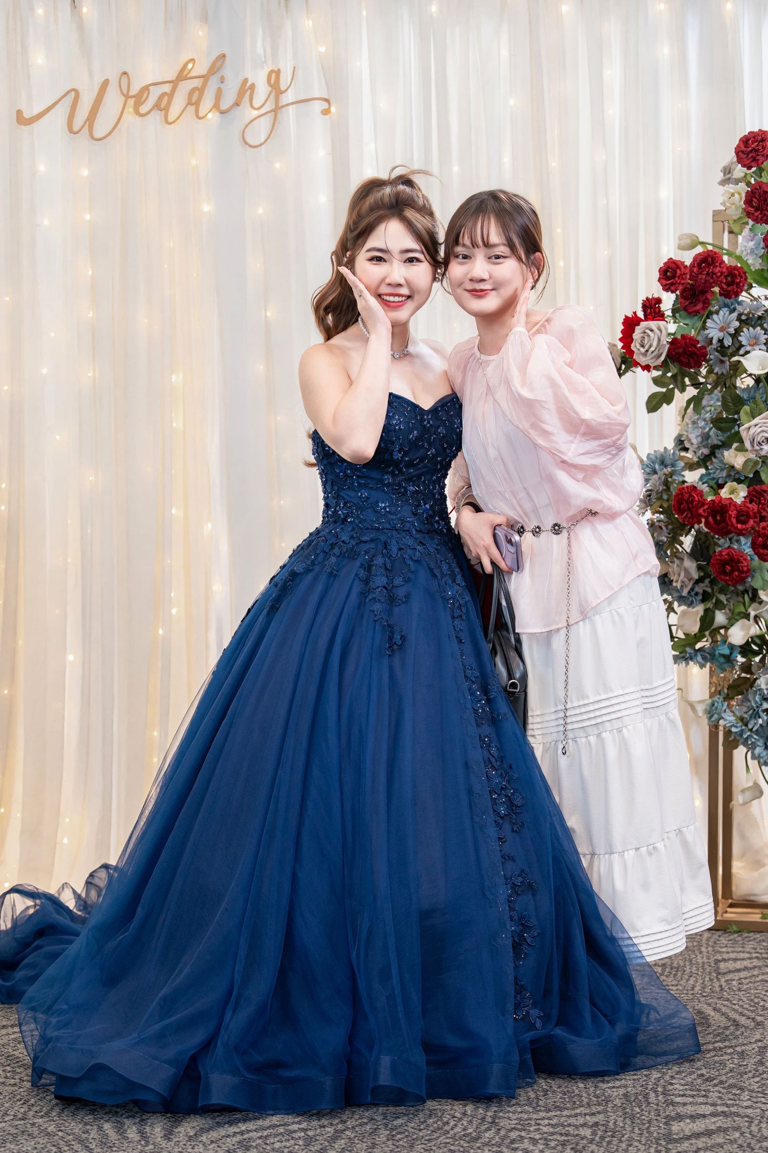 A person in a navy blue ball gown poses with others in white dresses against a light backdrop with red floral decor.