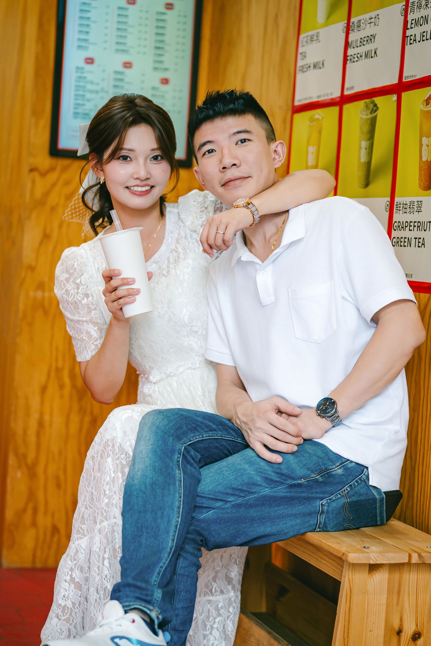 Couple in white dress and casual attire share drinks while sitting together against wooden wall with menu boards.