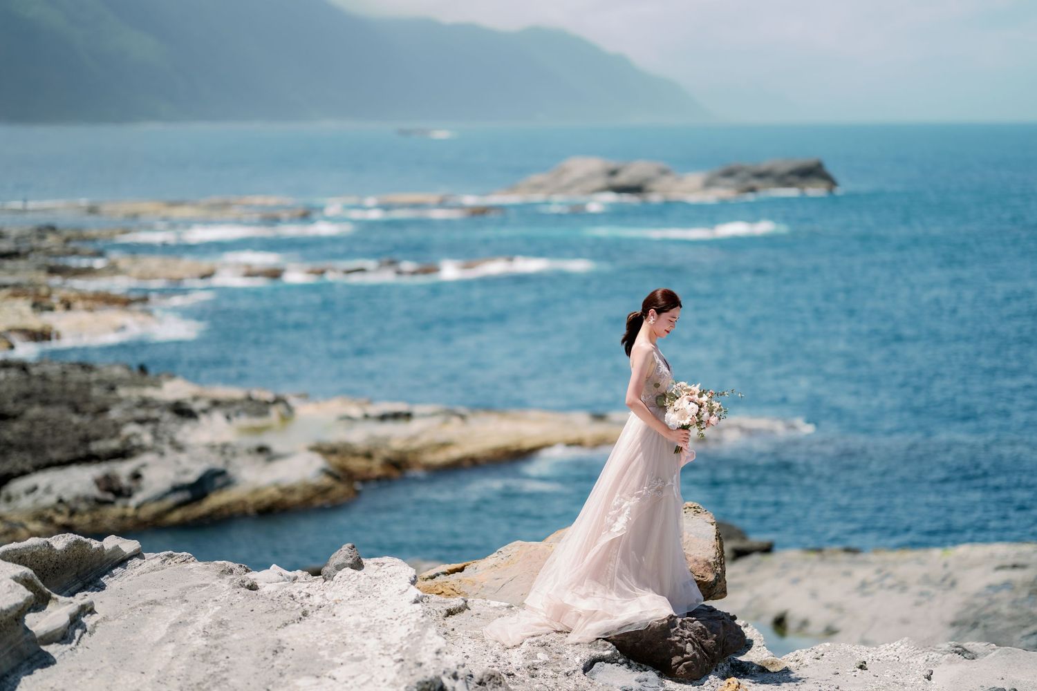 A bride in a white dress walks along rocky ocean cliffs as waves crash against the shore during a coastal photoshoot.