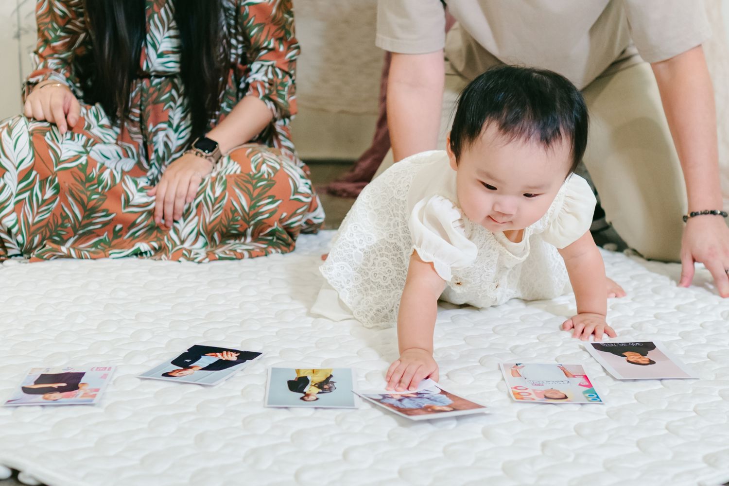 Baby in white dress crawls on blanket looking at playing cards spread out on the floor.