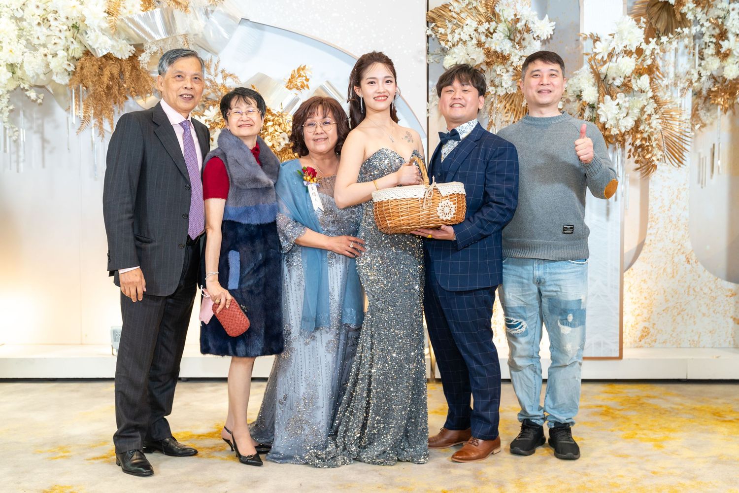 A group stands together in formal attire holding a decorative basket against an elegant backdrop with dried floral arrangements.