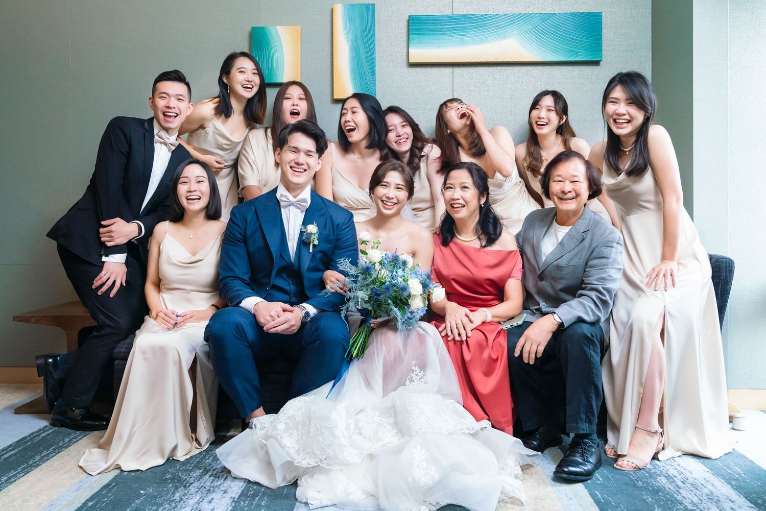 Wedding party dressed in formal attire poses together on couch at indoor venue with blue wall.