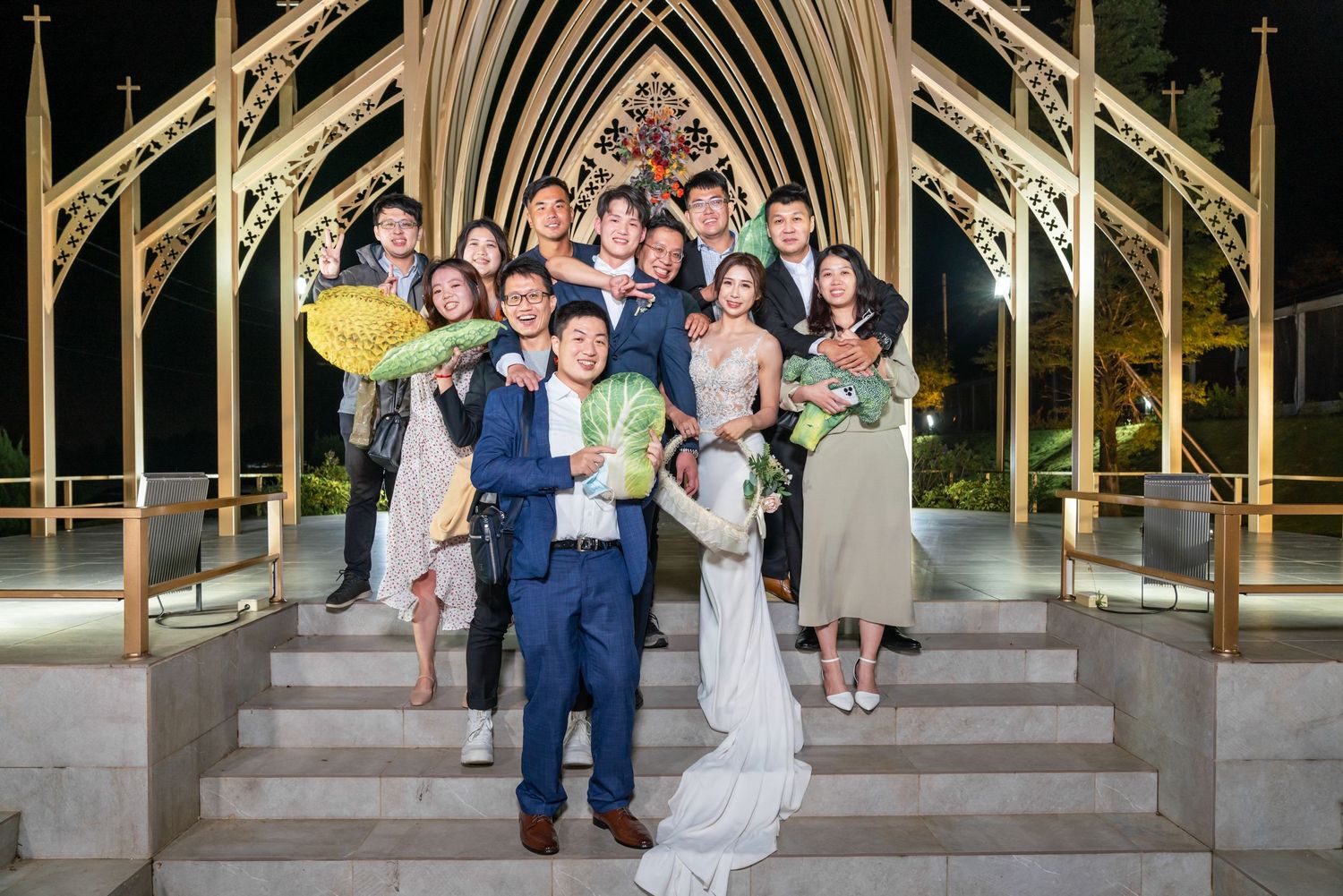 A wedding party poses on stone steps beneath an ornate golden Gothic archway at an outdoor chapel.