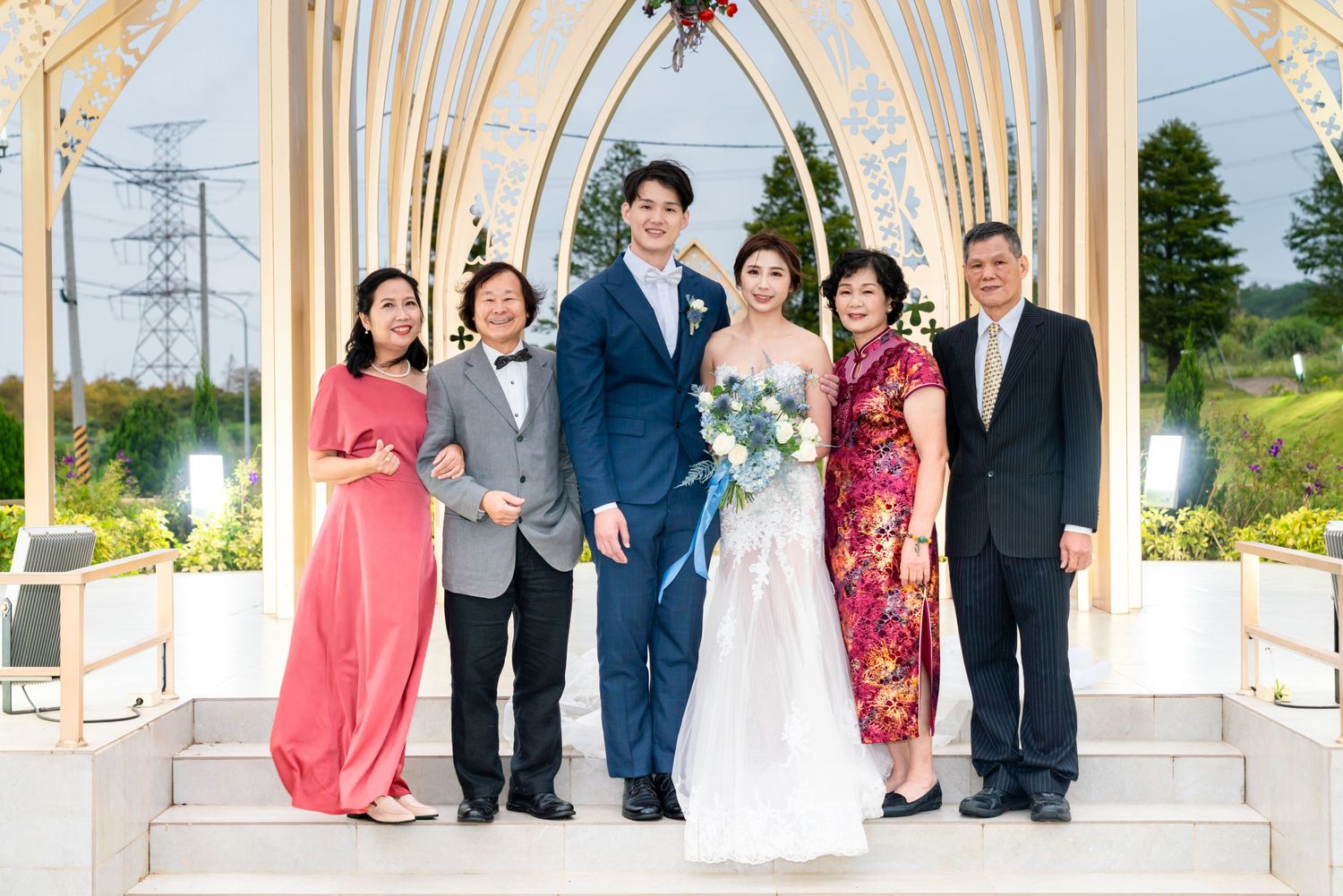 A wedding party poses together under an elegant white archway at an outdoor chapel venue on a sunny day.