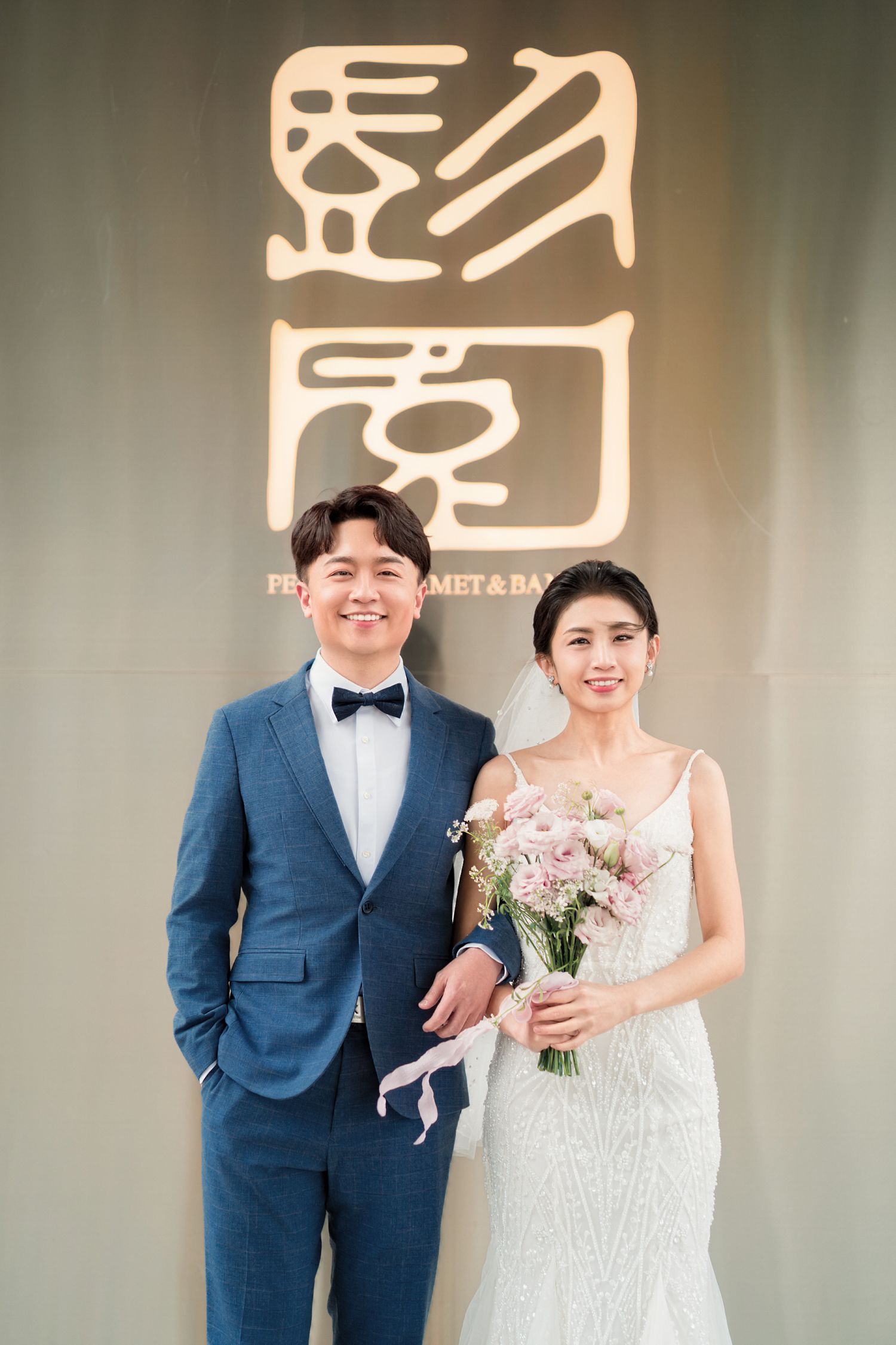 A formal couple poses together in wedding attire against a wall with Chinese characters at an upscale venue.