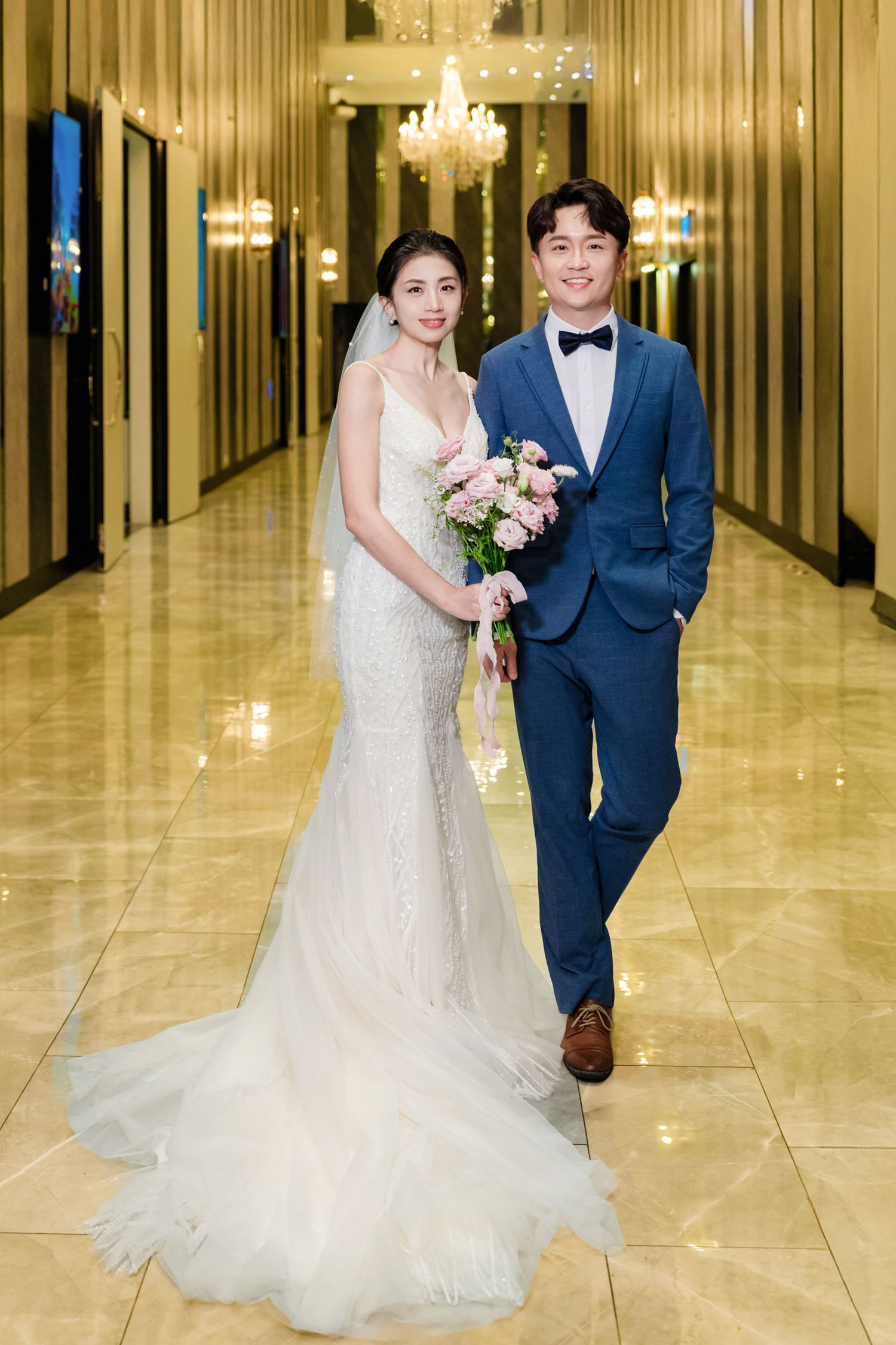 A newlywed couple poses in an elegant hallway with chandeliers, the bride in a flowing white gown holding a bouquet.