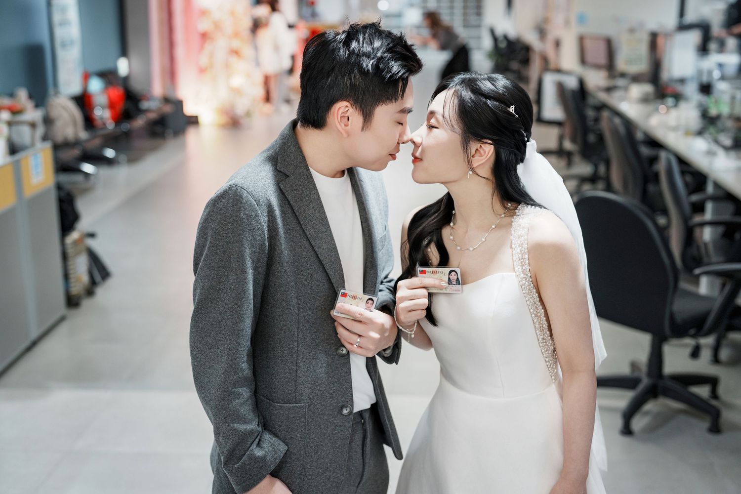 A romantic couple in wedding attire share an intimate moment in an office setting with computers and desks in background.